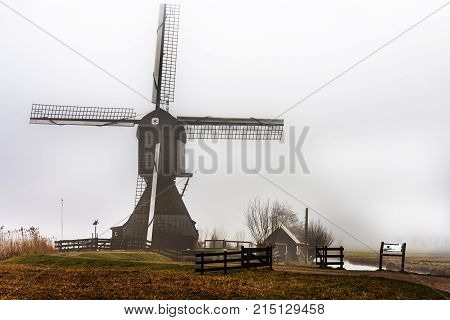 Silhouette Of A Dutch Windmill Under A Misty And Foggy Weather A Long A Calm And Quiet Canal
