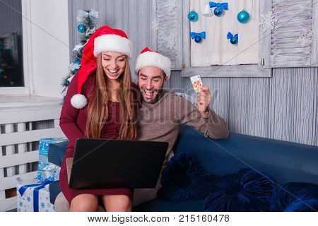 Happy young couple smiling, doing online shopping, on New Year's Eve, sitting on sofa at home, using credit card and laptop, the little one is sitting on his lap beside the man.