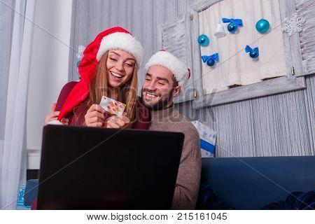 Happy young couple doing online shopping, on New Year's Eve, sitting on sofa at home, using credit card and laptop.