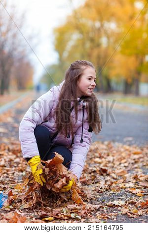 A girl in a pink jacket, sitting in an autumn park, collects the fallen leaves.