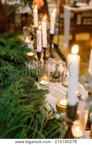 new year, interior design, winter concept. long table for celebrating decorated with numerous of shining candles in tall holders and in faceted thick wall flasses, they are standing by green branches