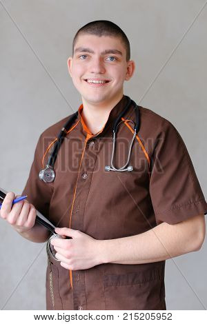 Handsome male specialized doctor poses on camera in good mood and smiles, loves work and gives advice standing near couch with folder and centimeter tape in his hands, and stethoscope on neck. 