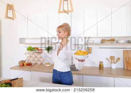 Attractive young woman presents dinner in plates, holds flat plate with tomatoes, cucumbers and lettuce leaves and deep plate with potato chips in palms, smiling with beautiful smile, standing in stylish light kitchen against backdrop of cabinets