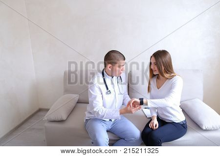 Successful male cardiologist who shows female on iWatch gadget, which girl on arm has to control analysis and adjustment of day's routine. Therapist with stethoscope and patient sitting on couch on background of gray wall in hospital's office during day. 