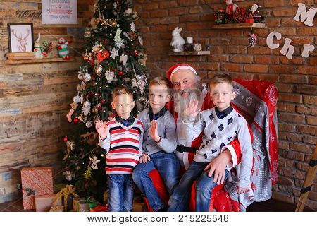 Merry brothers boys sitting on Santa Clauss lap and waving their palms camera rejoice. Claus with gray long beard dressed in red suit white trim pointed cap pompon end. Charming men of European appearance warm sweaters blue jeans socks, sit large soft arm