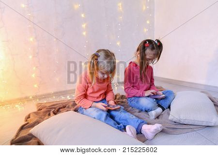 Lovely little girls holding mobile phones and playing games or taking pictures,  typing text for messages on touch screen, they laughing and smiling, sitting on rug in bright room with flickering New Year's garland in evening. Blond girl of European appea
