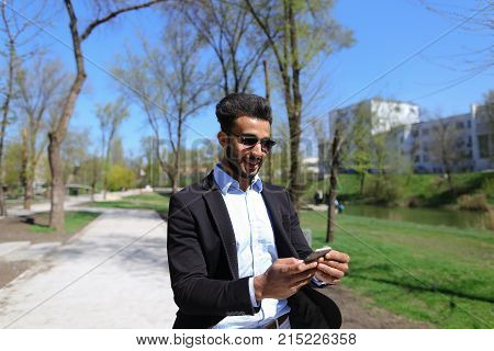 Man hold phone in hand and talking with mom. Son has dimple on cheeks, beard, short hair and long nose. Person dressed in black jacket and blue shirt. Concept of new apps modern technologies and long talks and cheep tariff plans.