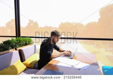 Freelance copywriter typing text fast on laptop keyboard at cafe table and looking tired. Handsome young man dressed in black shirt has dark hair and beard. Concept of typewriting skills and independent contractor for agencies.