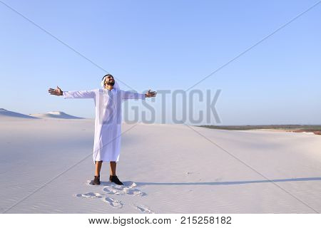 Stately young sheikh walks on foot along bottomless desert leaving traces on sand, enjoying warmth of day. Attractive Muslim smiles widely and spreads hands to sides, straightens clothes in wind and whirls around, delighted by breadth of desert. Swarthy, 