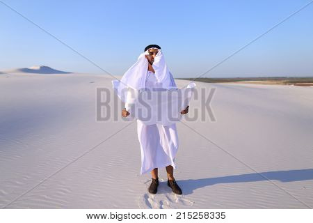 Beautiful Emirate Sheikh businessman studying project of future construction of complex, standing in middle of bottomless desert with white clean sand on clear warm day against blue sky. Swarthy Muslim with short dark hair dressed in kandura, long, spacio