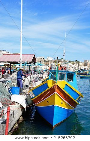 MARSAXLOKK, MALTA - APRIL 1, 2017 - Traditional Maltese Dghajsa fishing boats in the harbour with waterfront buildings to the rear and a local man sorting his fishing nets on the quayside Marsaxlokk Malta Europe, April 1, 2017.