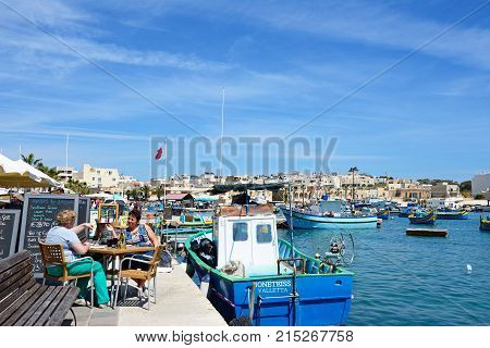 MARSAXLOKK, MALTA - APRIL 1, 2017 - Traditional Maltese Dghajsa fishing boats in the harbour with waterfront buildings to the rear and two women relaxing at a restaurant on the quayside Marsaxlokk Malta Europe, April 1, 2017.