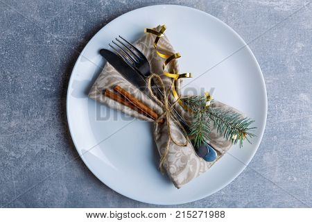 On a worn surface, a round white plate with serving cutlery is decorated with a fir tree branch, cinnamon sticks and a golden ribbon. Christmas atmosphere. View from above. Indoors.