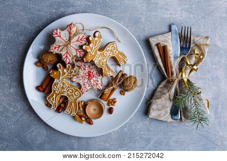 In a round white plate a lot of different cookies of different shapes with sugar icing, next to them there are cutlery wrapped in a tissue napkin decorated with a Christmas tree branch, cinnamon sticks and a golden ribbon. Christmas atmosphere. Indoors.