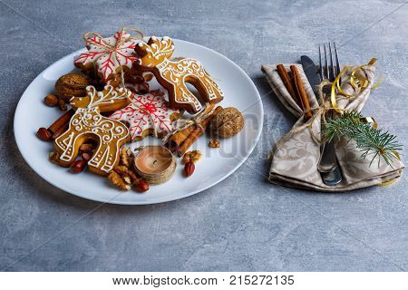 On the shabby surface a white round plate with tasty Christmas cookies of different shapes with sugar icing, next to there are cutlery wrapped in a tissue napkin and decorated with a Christmas tree branch, cinnamon sticks and golden ribbon.