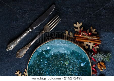Round plate in dark glossy surface, next to old cutlery, wooden snowflakes, Christmas tree branch and cinnamon sticks. Close-up. Indoors.