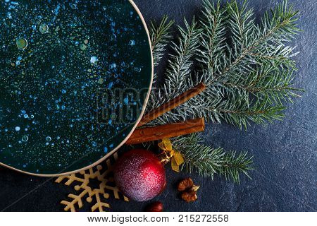 On the table, a delicious plate in dark glossy surface and a Christmas tree branch is decorated with a ball and cinnamon sticks. View from above. Close-up. Indoors.