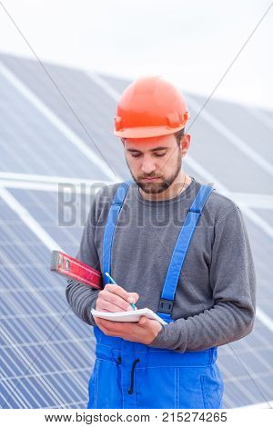 A solar station worker in blue uniform and in an orange helmet, holds a waterpas in his hands and writes something into a notepad.