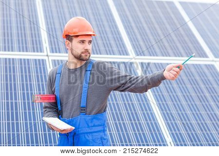 A young worker stands a solar cell station in uniform and a helmet holds a waterpas and a notebook in his hands and shows a hand somewhere. Outdoors.