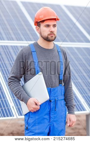 An employee of the solar battery station with a beard, dressed in uniform and a helmet stands and looks somewhere with a serious look, holding a gray modern laptop in his hands. Outdoors.