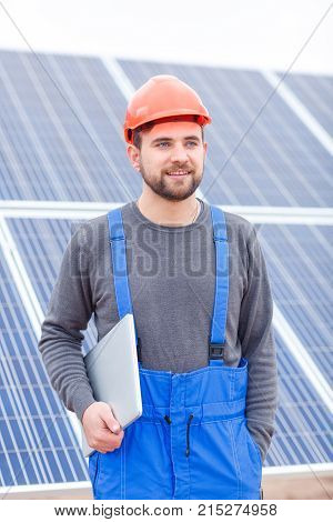 Stations of solar battery. The worker in blue uniform and in an orange helmet stands near the panel and holds a modern laptop in his hands and looks somewhere far away. Outdoors.