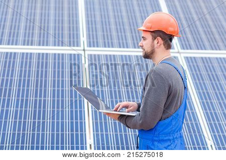 A male worker in blue uniform and an orange helmet stands near the panel and holds an open modern laptop in his hands and looks somewhere. Outdoors.