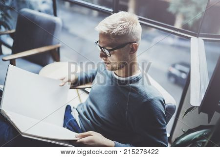 Portrait handsome young man wearing glasses casual clothes holding book hands.Man sitting in vintage armchair modern loft studio reading book. Blurred background.Horizontal