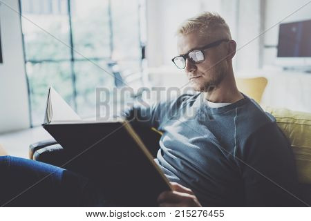 Portrait handsome young man wearing glasses casual clothes holding book hands.Man sitting in vintage armchair modern loft studio reading book. Blurred background.Horizontal