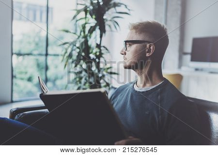 Portrait handsome young man wearing glasses casual clothes holding book hands.Man sitting in vintage armchair modern loft studio reading book. Blurred background.Horizontal