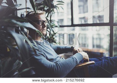 Attractive young man wearing glasses casual clothes.Man sitting in vintage armchair modern loft studio and relaxing whith headphone music. Blurred background.Horizontal