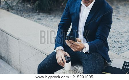 Attractive smiling businessman using contemporary smartphone taking rest during work day.Holding hands take away coffee cup.Blurred background. Horizontal.Cropped