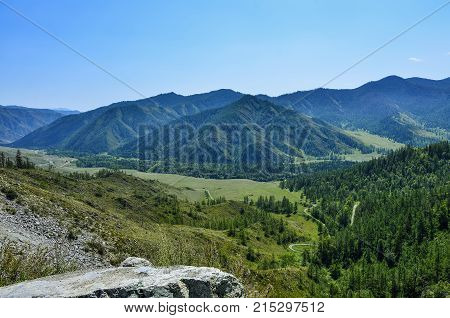 Beautiful summer mountains landscape Altai Russia. Mountain ridges covered with forest a road through the valley and large boulder at foreground