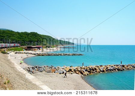 Summer. Blue sea. Blue sky. The sun is shining. The shore of the sea with breakwaters made of huge stones. Along the coast there is a railway on which the train is going. In the distance you can see the mountains.