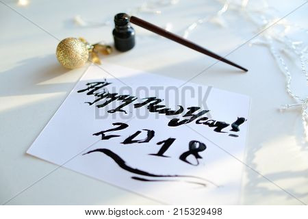 Christmas card with festive inscription Happy New Year, painted with black calligraphic font using carcass and fountain pen, which lies on table with Christmas garland in art workshop. Concept of beautiful writing, calligraphy and lettering, materials and