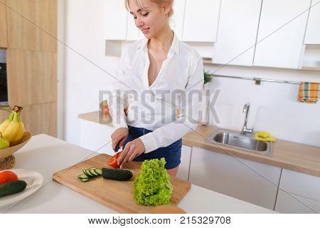 Attractive young woman cuts green cucumber for salad with sharp knife, takes red juicy tomato and several times throws up with smile on face, sniffs freshness of lettuce leaves and takes up shredding vegetables. Blonde at top of modern light kitchen with 
