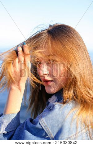 Attractive young woman with smile on face turns head to sides and develops beautiful hair, turns in profile and poses, holding hands on belt and stands with back to camera lens on sea shore against blue sky on clear summer day. Woman of European appearanc