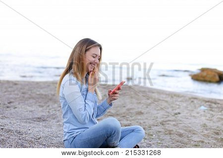 Cute girl holding cell phone and leads in social network or post photos, laughing and smiling, sharing impressions of beauty of sea, sitting on sandy beach on backdrop of sea and sky on clear evening . Young European-looking woman with long blond hair dre