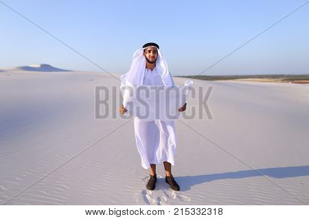 Beautiful Emirate Sheikh businessman studying project of future construction of complex, standing in middle of bottomless desert with white clean sand on clear warm day against blue sky. Swarthy Muslim with short dark hair dressed in kandura, long, spacio