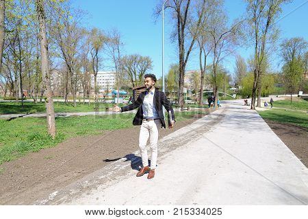Guy walking in park and smiling, slow motion. Man dressed in blue shirt, white jeans and brown shoes. Boy has black hair and beard. Concept of places for rest and clean parks and cheeps fashion clothes for man