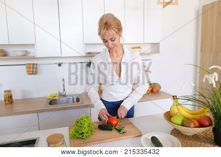 Attractive young woman cuts green cucumber for salad with sharp knife, takes red juicy tomato and several times throws up with smile on face, sniffs freshness of lettuce leaves and takes up shredding vegetables. Blonde at top of modern light kitchen with 