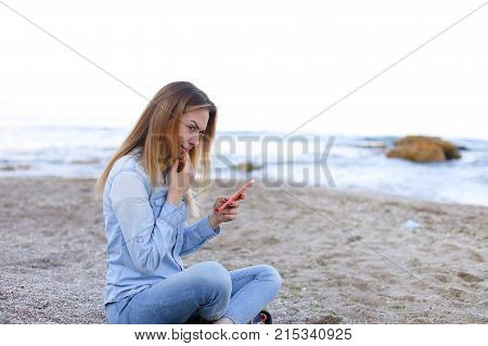 Cute girl holding cell phone and leads in social network or post photos, laughing and smiling, sharing impressions of beauty of sea, sitting on sandy beach on backdrop of sea and sky on clear evening . Young European-looking woman with long blond hair dre