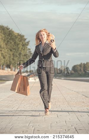 Vertical orange tonted shot of a happy beautiful blond woman with braces running with paper bags and phone in hand along the promenade