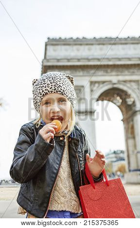 Stylish autumn in Paris. Portrait of smiling elegant child with red present bag near Arc de Triomphe in Paris eating French macaroon