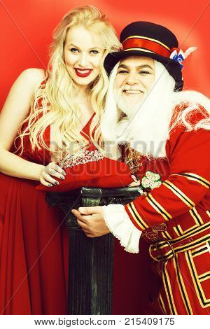 Happy senior man beefeater yeomen warder in uniform and pretty girl young sexy blond woman in dress with crown smile on red wall