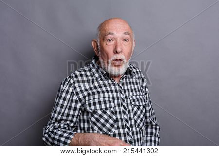 Surprised senior man in casual checkered shirt grimacing at gray studio background with copy space. Shok and excitement concept