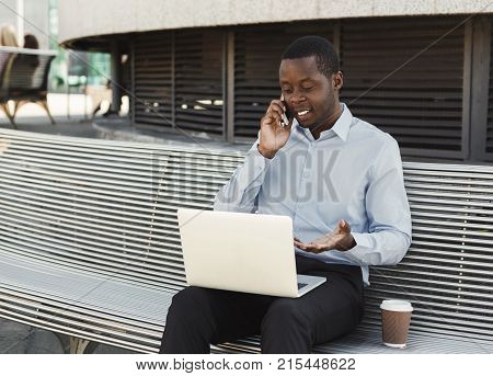 Happy black businessman working on laptop. African-american insuranse agent checking email on computer, talking on mobile, sitting on the bench outdoors near modern office center
