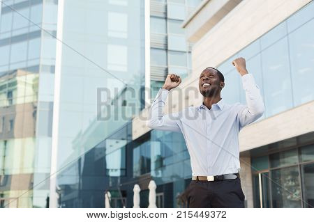 Happy excited black businessman celebrate victory with fists raised in the air. Winner, successful african-american man outdoors against modern glass office center background, lifestyle portrait