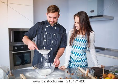 A young couple in the kitchen, the man smiles, sifts the flour, all in flour, the little girl watches.