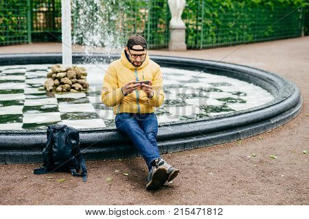 Young Attractive Bearded Man In Casual Clothes Sitting Near Stone Fountain Being Concentrated Into S