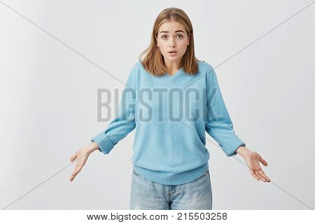 It's not a big deal. Clueless cute girl dressed in blue sweater and jeans shrugging shoulders and staring at camera with confused look after she did something wrong but not feeling sorry or guilty for that.